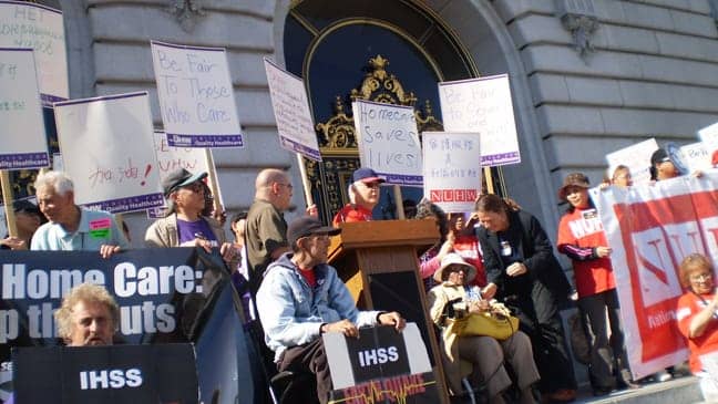IHSS-Earthquake-Geraldine-Holloway-with-mic-intro’d-by-Cathy-Spensely-dir.-Fam-Svcs-Agency-Bruce-Allison-sits-with-sign-102209-SF-City-Hall, IHSS Earthquake: Seniors fight for In-Home Supportive Services, Local News & Views 